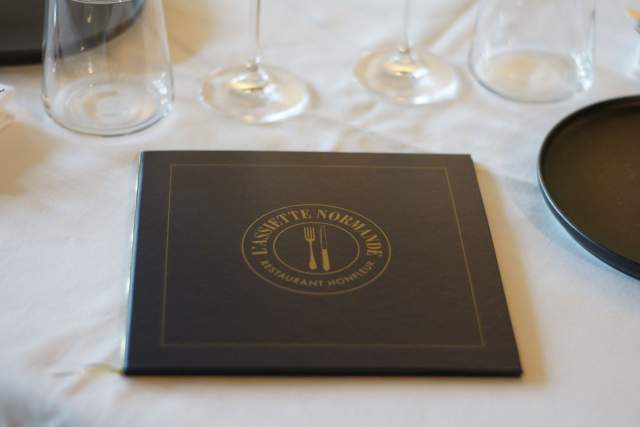 Carte du restaurant L'Assiette Normande en cuir noir posée entre deux verres à pied, restaurant L'Assiette Normande à Honfleur